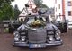 Brautpaar jubelt aus einem Oldtimer mit Blumenschmuck vor historischer Kulisse.