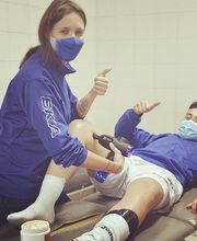 Persona aplica terapia en la pierna de un hombre, ambos con mascarillas y ropa azul.