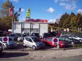 Gebrauchtwagen auf Parkplatz vor einer Autowerkstatt, umgeben von Bäumen, Himmel blau mit Wolken.