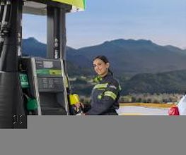 Una mujer en uniforme junto a un surtidor de gasolina con montañas al fondo.