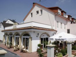 Weißes Gebäude mit Restaurant, Außensitzbereich, Krombacher-Schirm und blauer Himmel im Hintergrund.