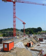 Baugelände mit zwei roten Kränen und Containern, umgeben von Bäumen unter blauem Himmel.