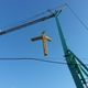 Una croce di legno viene sollevata da una gru contro un cielo blu.