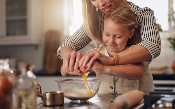 Mutter backt mit ihrer Tochter. Sie schlagen zusammen ein Ei über einer glutenfreien Mehlmischung in einer Glasschüssel auf.