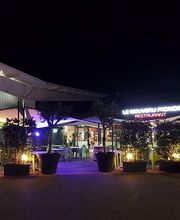 Nachtaufnahme eines beleuchteten Restaurants mit Bäumen in großen Pflanzkübeln davor.