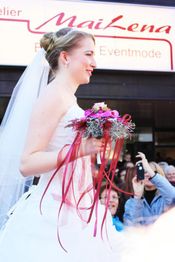 Braut mit Blumenschmuck in der Hand, lächelnd vor einem Geschäft für Braut- und Eventmode.