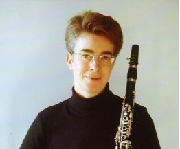 Person mit Kurzhaarfrisur hält eine Klarinette, trägt schwarzen Rollkragen und Brille.