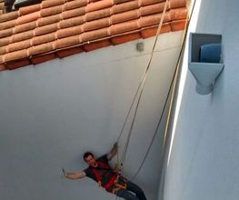 Un obrero colgado en unas cuerdas sonríe y toma una foto en una fachada blanca.