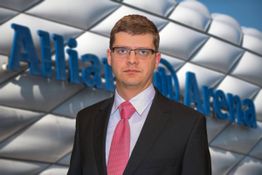 Ein Mann im Anzug steht vor einem Schild der Allianz Arena. Hintergrund: moderne Architektur.