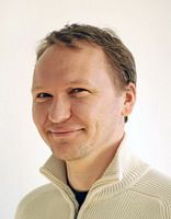 Ein Mann mit hellem Pullover lächelt vor neutralem Hintergrund.