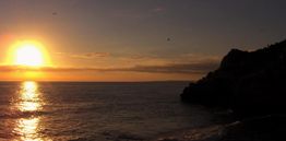 Sonnenuntergang über dem Meer mit einer dunklen Felsenküste rechts; goldene Reflexionen im Wasser.