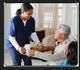 Caregiver serving food and drink to an elderly man seated with a child in a cozy living room.