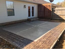 Concrete patio with decorative brick border in a fenced backyard by a house.