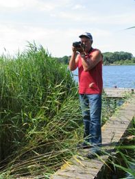 Mann fotografiert auf einem Steg am Seeufer mit hohem Schilf und klarem Himmel im Hintergrund.