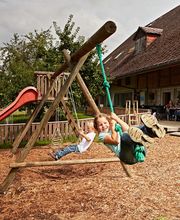 Kinder schaukeln fröhlich auf einem Spielplatz, im Hintergrund ein Haus mit Menschen auf der Terrasse.