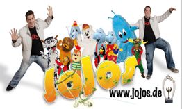 Zwei Männer posieren neben bunten Tierkostümen und dem Schriftzug "Jojos", Website-Link enthalten.