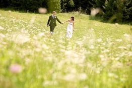 Paar in Hochzeitskleidung geht über eine blühende Wiese, von Bäumen im Hintergrund umgeben.