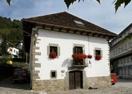 Casa antigua de paredes blancas, con balcones y flores rojas, rodeada de montañas y autos.