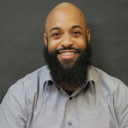 Smiling person with a beard and shaved head, wearing a striped shirt against a dark background.