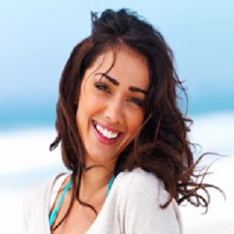 Smiling woman with wavy hair at the beach, wearing a light top, blue sky in the background.