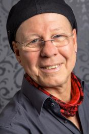 Mann mit Brille, Mütze und rotem Halstuch lächelt vor einem gemusterten Hintergrund.