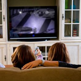 Two people sitting on a couch watching TV, one holding a remote.