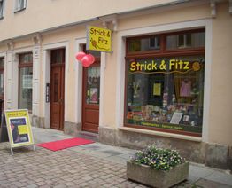 Fassade eines Handarbeitsladens "Strick & Fitz" mit roten Ballons und Blumen vor dem Eingang.