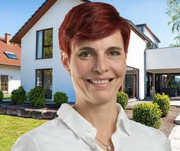 Eine lächelnde Frau mit kurzen roten Haaren steht vor einem modernen weißen Haus mit Garten.