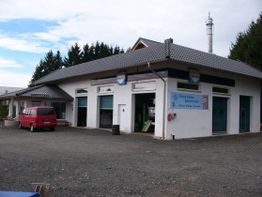 Weißes Gebäude mit grauem Dach, rotem Auto davor, umgeben von Wald, klarer Himmel.