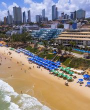 Praia movimentada com guarda-sóis coloridos, edifícios altos ao fundo e céu azul com nuvens.