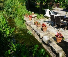 Jardín con mesa, sillas, y macetas junto a un estanque rodeado de vegetación verde.