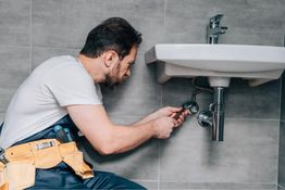 Homme réparant un tuyau sous un lavabo avec une clé, portant une ceinture à outils.