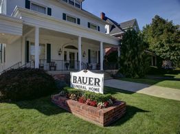 Funeral home with "Bauer Funeral Home" sign, surrounded by lawn and plants.