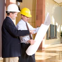 Two men in hard hats review architectural blueprints in a bright, modern lobby.