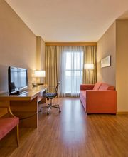 Sala de estar de hotel com sofá, mesa de trabalho, TV e vista para quarto com cama.