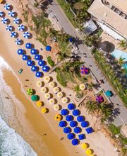 Vista aérea de uma praia com guarda-sóis coloridos, mar à esquerda e piscina à direita.