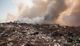 Burning landfill with smoke rising, surrounded by scattered waste under a hazy sky.