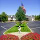 Parking lot with a flagpole, statue, and flowers leading to a building under a clear blue sky.
