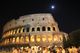 Colosseo illuminato di notte con la luna e folla di persone in movimento.