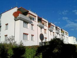 Mehrstöckiges Wohngebäude mit Balkonen, umgeben von grüner Hecke, unter klarem blauem Himmel.