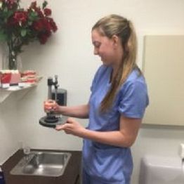 Smiling woman in scrubs holds a dental tool in a clinic room, with roses on a shelf nearby.