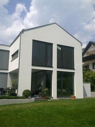 Modernes, weißes Haus mit großen Fenstern und Garten bei bewölktem Himmel.