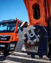 Drei orange-farbene Baufahrzeuge und ein Bagger mit "Powertilt"-Aufschrift bei sonnigem Wetter.