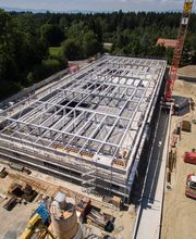 Bau einer großen Halle mit Stahlkonstruktion, umgeben von Baugeräten und Bäumen im Hintergrund.