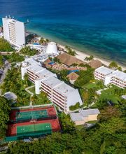 Vista aérea de un complejo hotelero junto al mar con canchas deportivas y vegetación abundante.