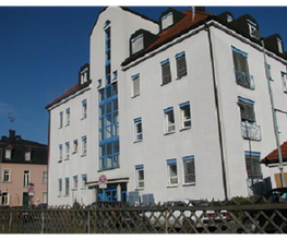 Mehrstöckiges Gebäude mit weißen Wänden und blauen Fensterrahmen, sonniger Himmel.