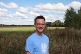 Mann im blauen T-Shirt steht auf einem Feld mit Bäumen im Hintergrund unter blauem Himmel mit Wolken.