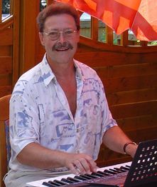 Mann mit Brille spielt Keyboard, trägt ein gemustertes Hemd, sitzt vor einer Holzwand.