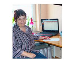 Person am Schreibtisch mit Laptop, trägt gestreiftes Hemd. Fenster, Pflanzen im Hintergrund.
