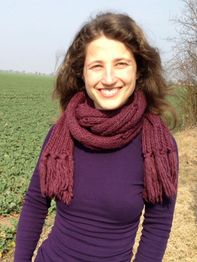 Frau mit lila Schal lächelt auf einem Feldweg vor einem grünen Feld bei sonnigem Wetter.